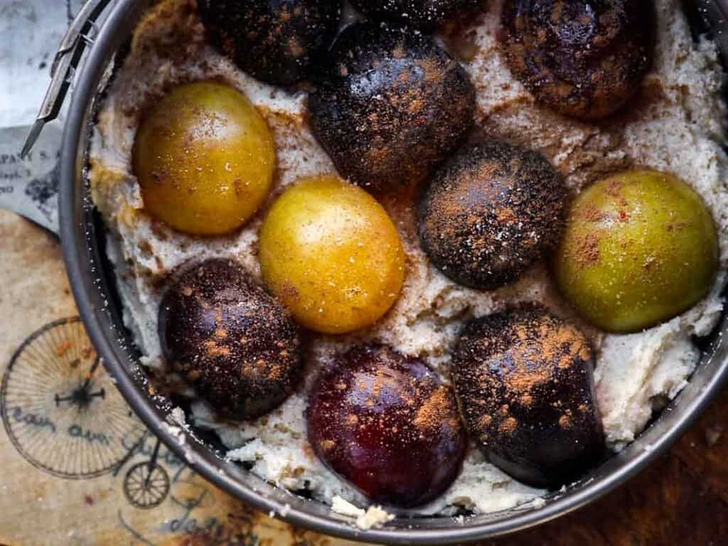 A close-up of a baking pan filled with a creamy batter topped with whole plums of varying colors, dusted with cinnamon, ready to be baked. The pan rests on a brown paper with bicycle illustrations.
